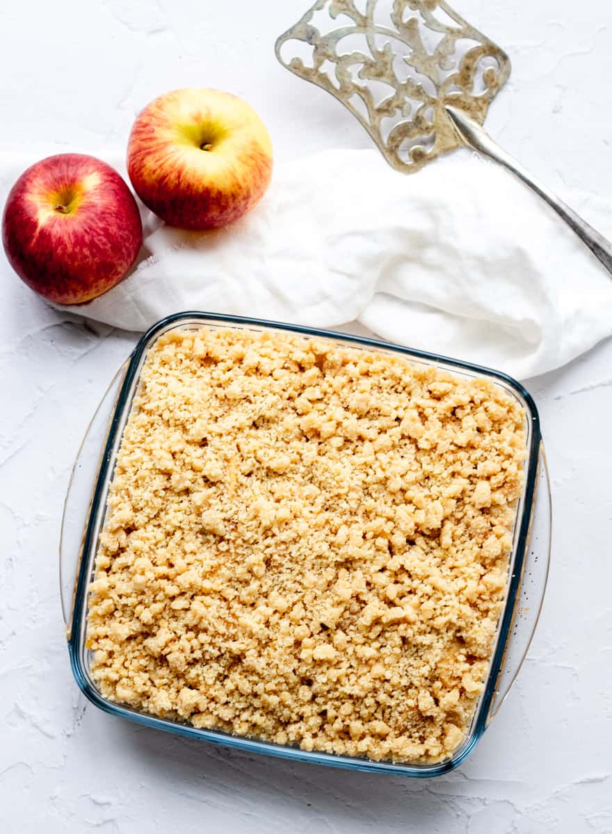 Glass dish of apple crumble with apples, cloth and serving spatula on grey background.