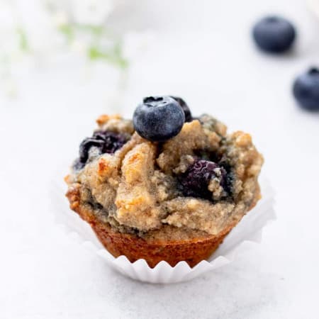a mini almond flour blueberry muffin in a white paper liner with fresh blueberry on top
