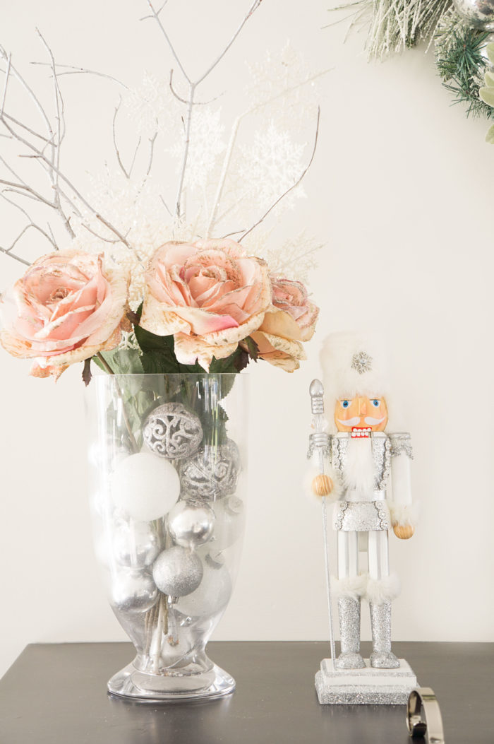 White and silvery nutcracker next to a vase with pink flowers and silver bulbs