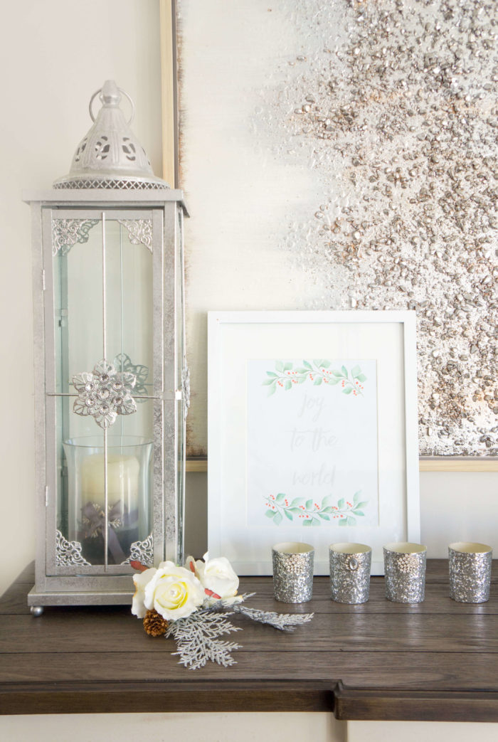 Framed paper that reads \"Joy to the World\" next to a tall silver lantern and silver candle holders