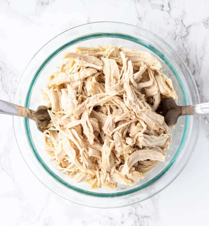Shredded chicken in a glass bowl with two forks on a marble background