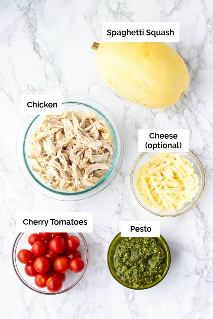 Ingredients for pesto chicken stuffed spaghetti squash boats on a marble background with labels