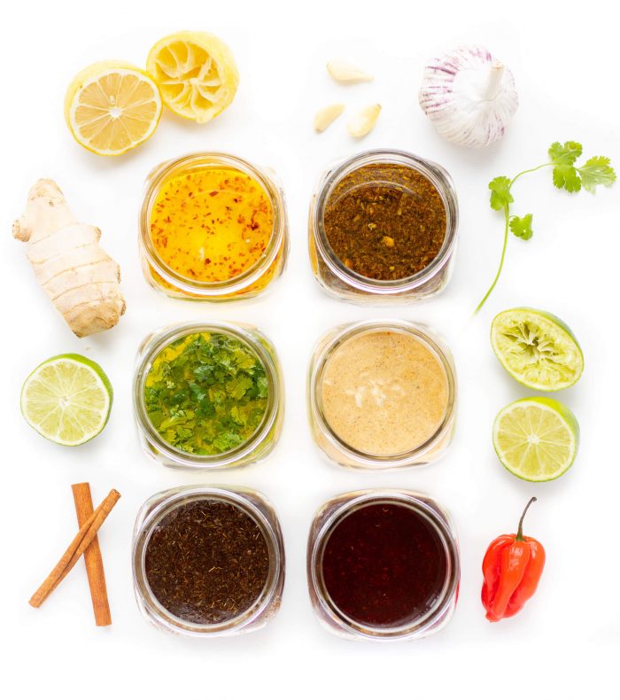 Six open jars of marinade surrounded by ingredients including lime, ginger, cilantro, and garlic