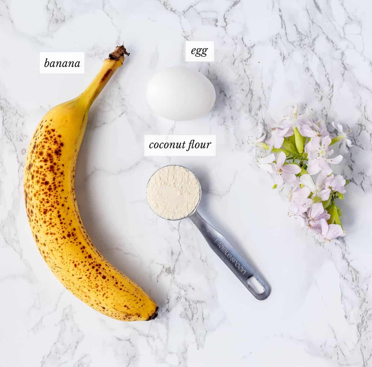 ingredients for banana egg pancakes with labels next to some pink flowers on a marble background