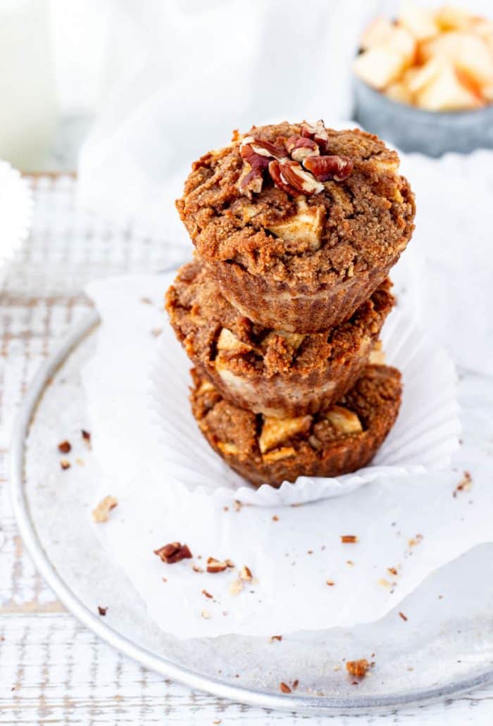 three apple muffins stacked with chopped pecans sprinkled around with chopped apple in background