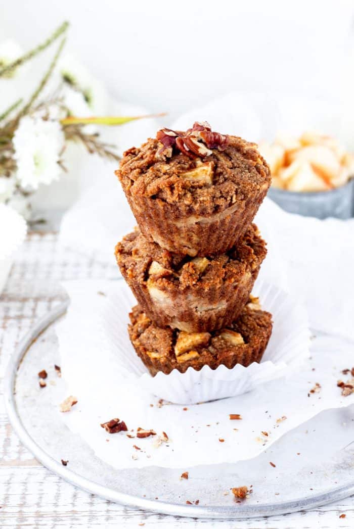 three apple muffins stacked with chopped pecans sprinkled around with apples and flowers in background