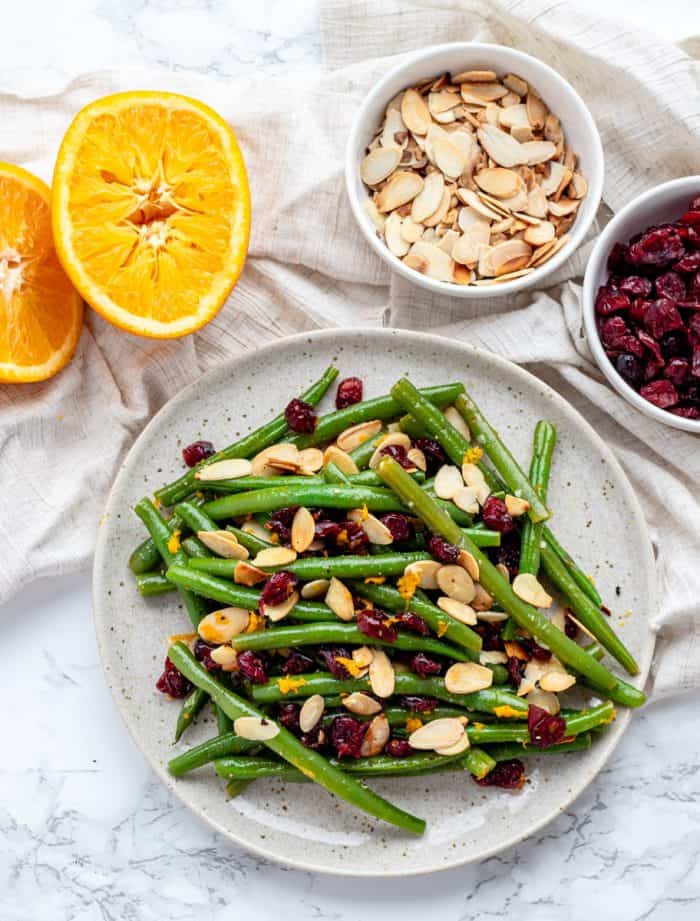 green beans with garlic, dried cranberries, almonds and orange zest on plate with orange and almonds in background