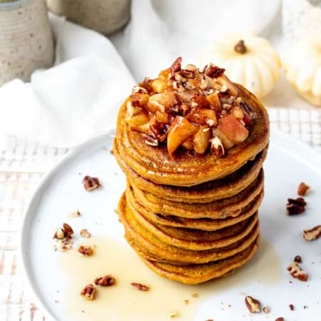 stack of pumpkin pancakes on white plate with a sprinkle of pecans and apple topping with white pumpkins in background