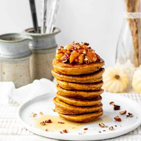 stack of pumpkin pancakes on white plate with a sprinkle of pecans and apple topping with white pumpkins in background