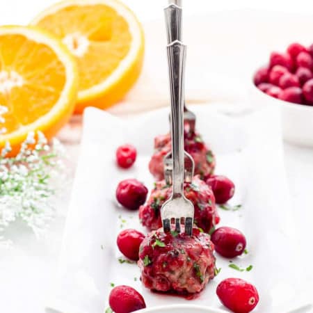 Cranberry meatballs on a white serving plate with small forks in them on a white tray with an orange and cranberries in background.