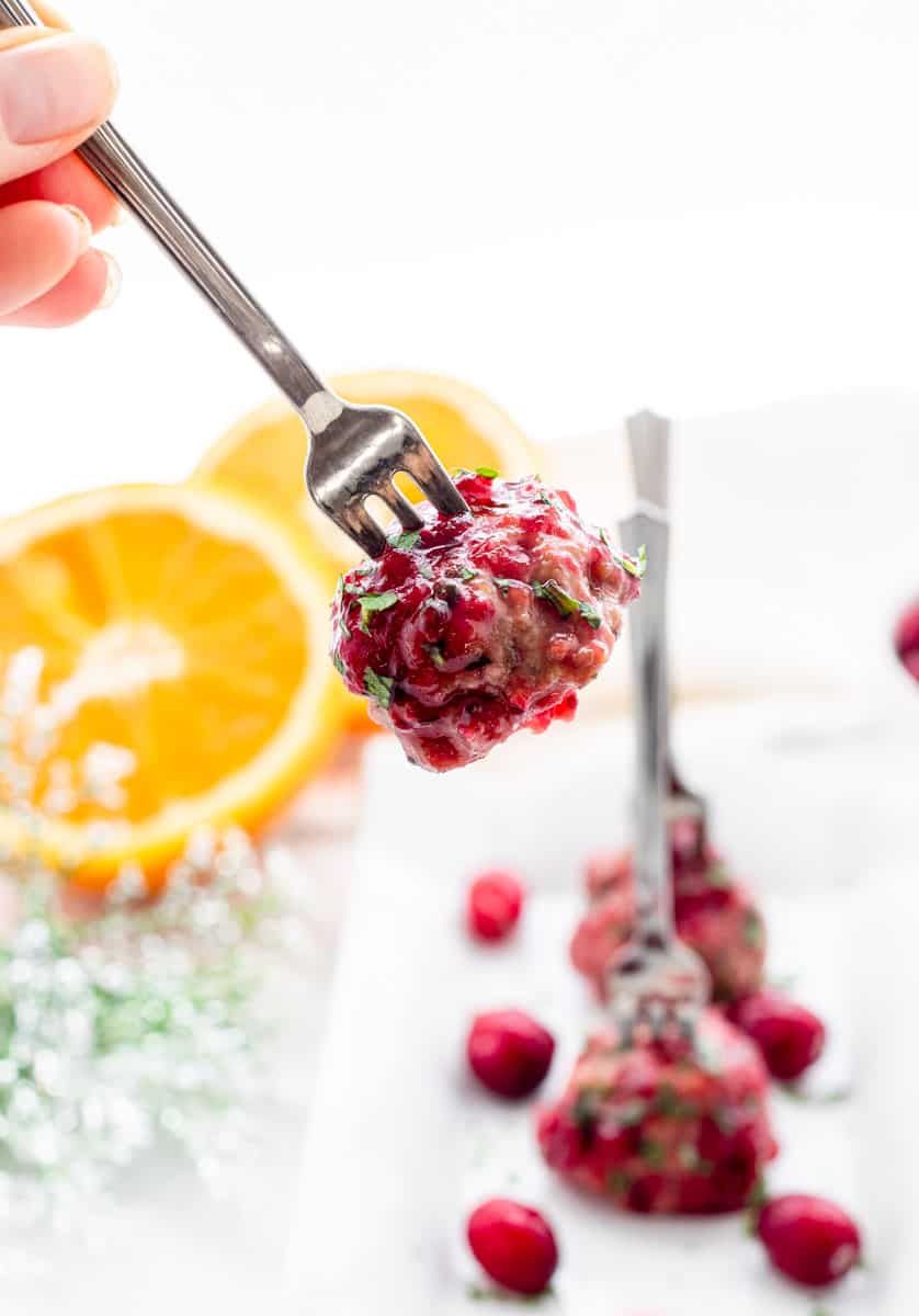 A cranberry party meatball on a small fork with meatballs on white tray next to an orange.