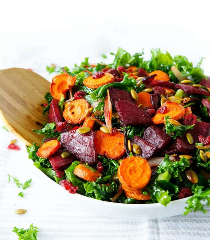 Roasted beets and carrots on a bed of kale in a white bowl with a wooden serving spoon.