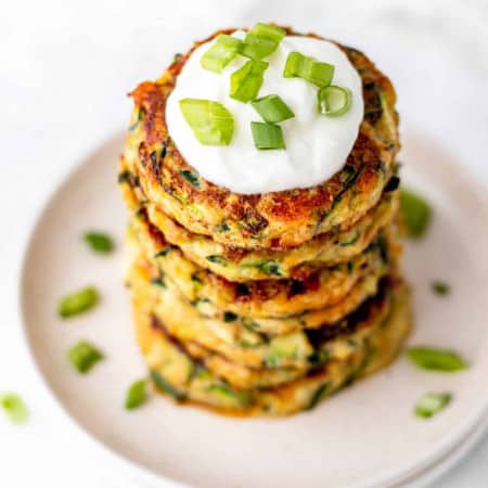 Zucchini fritters stacked on a white plate with a dollop of sour cream.