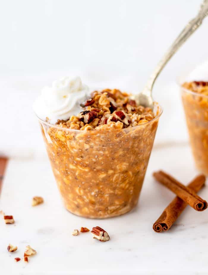 A spoon in a cup of pumpkin overnight oats next to cinnamon sticks.