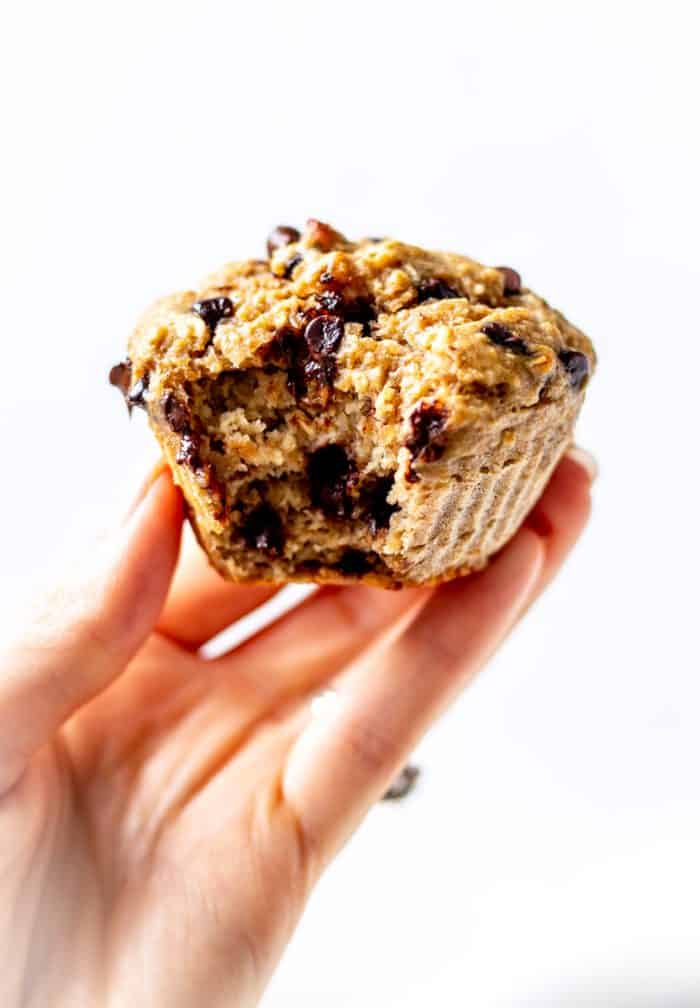 A hand holding a banana protein muffin with a bite taken out of it.