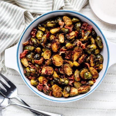 overhead shot of roasted brussel sprouts and bacon in white and bowl bowl next to two large serving forks