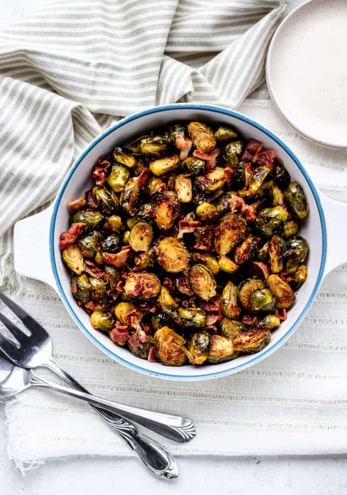overhead shot of roasted brussel sprouts and bacon in white and bowl bowl next to two large serving forks