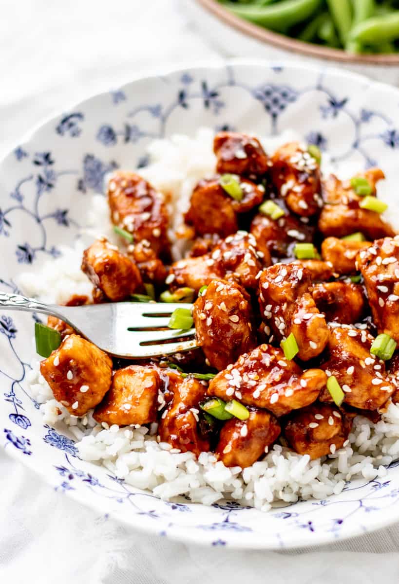 fork piercing orange chicken on a bed of rice in a blue and white bowl