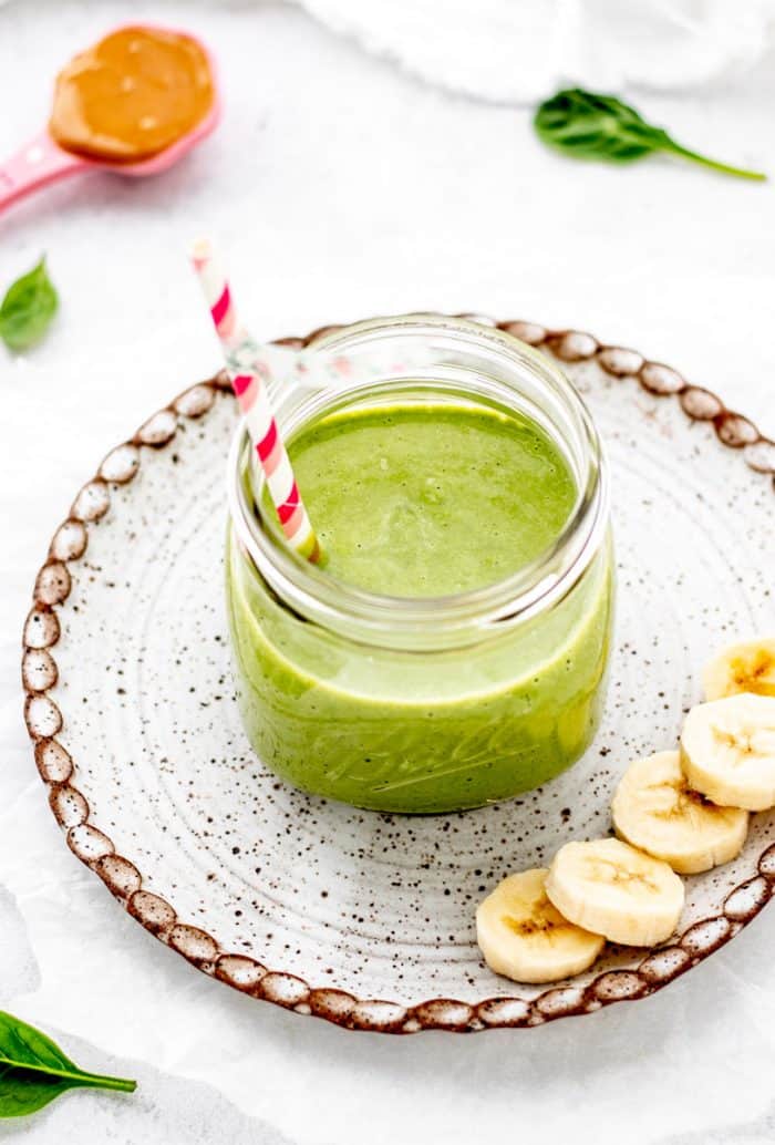green monster smoothie in a mason jar on a plate with sliced banana and spinach.
