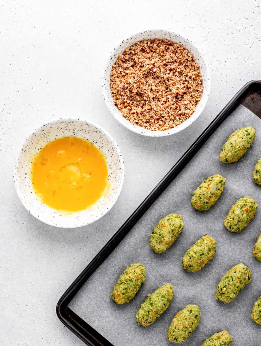 Broccoli tots on a baking sheet next to bowls of egg and bread crumbs.