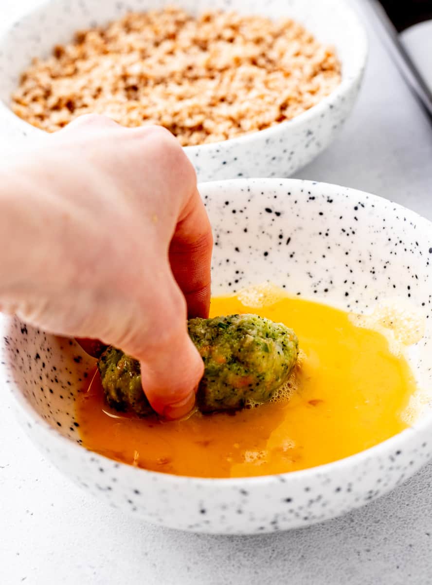 A hand dipping broccoli tot in a bowl of beaten egg.