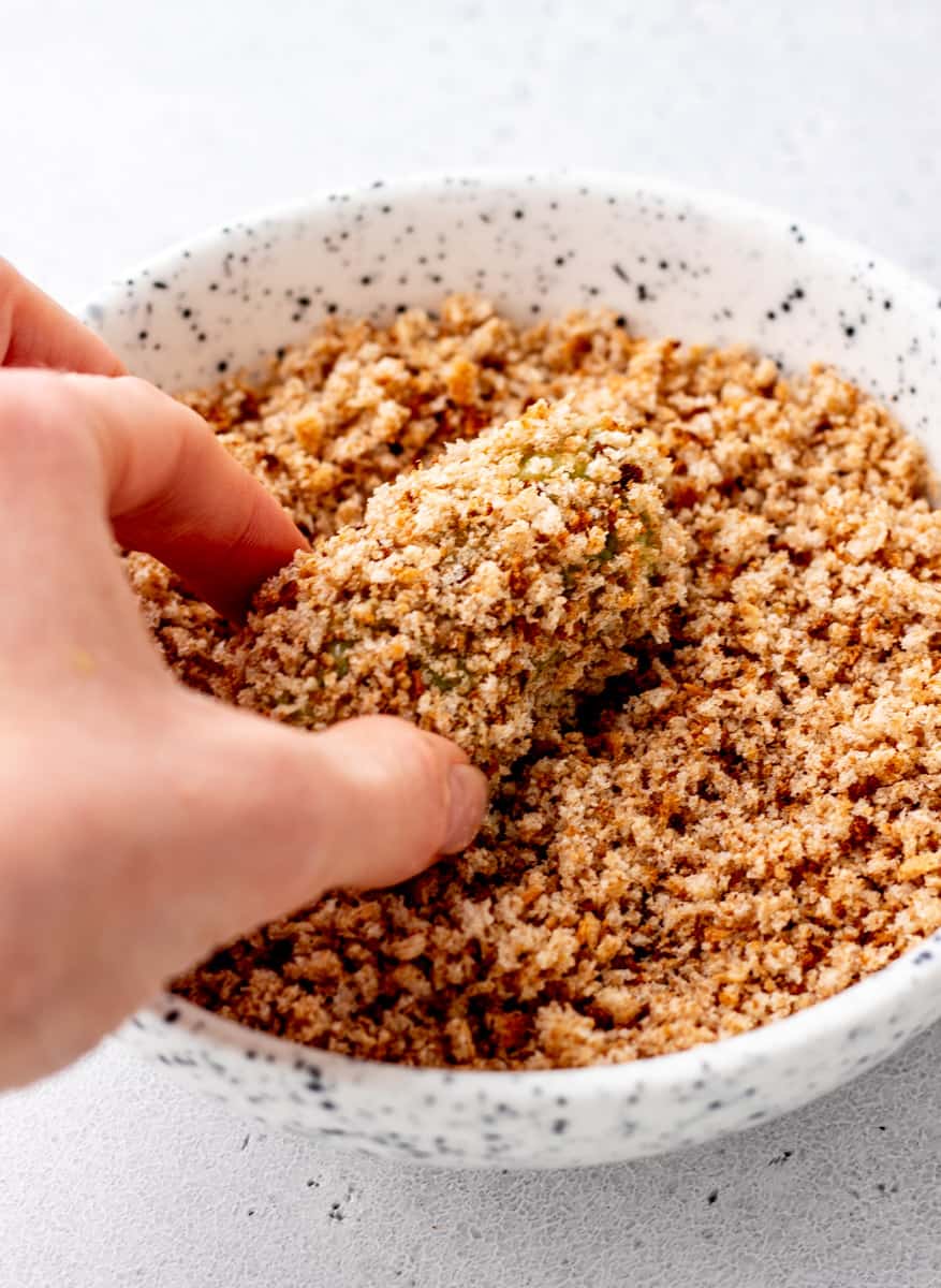 A hand dipping broccoli tot in a bowl of bread crumbs.