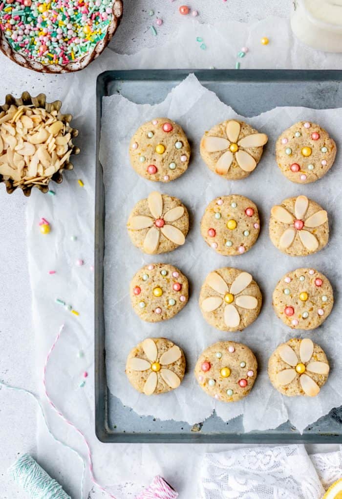 Decorated vegan lemon cookies on a tray next to almonds and sprinkles.