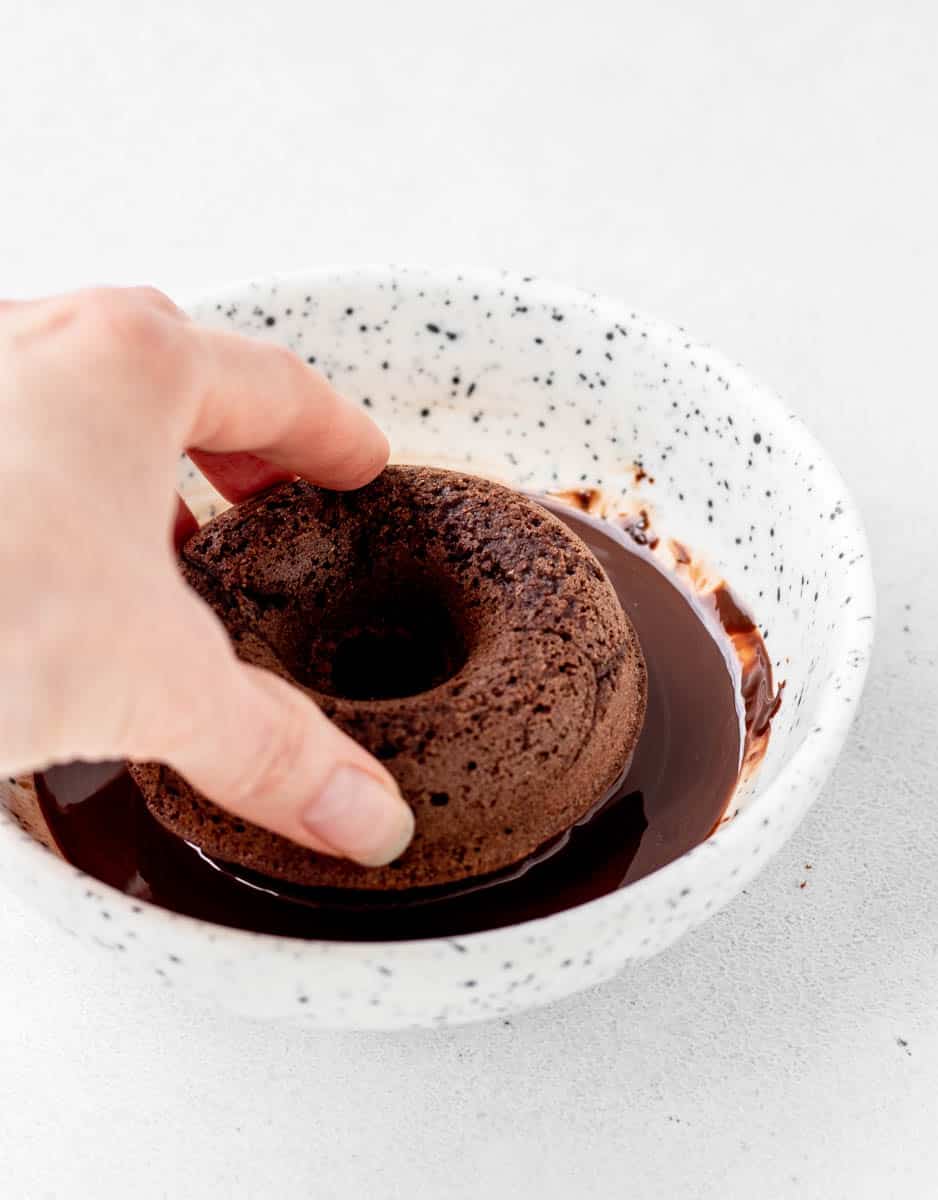 A hand dipping a chocolate donut into chocolate glaze.