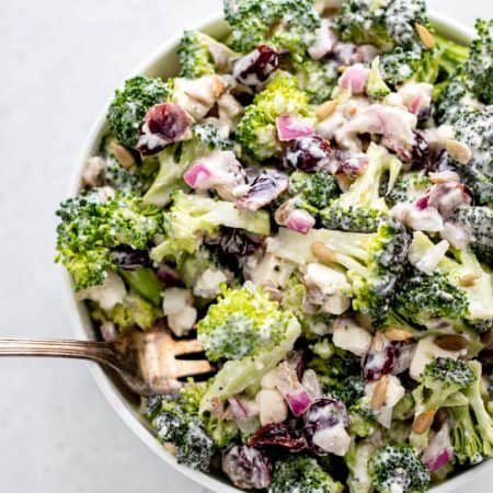 A fork in a bowl of broccoli salad with sunflower seeds, cranberries, red onion and creamy dressing.