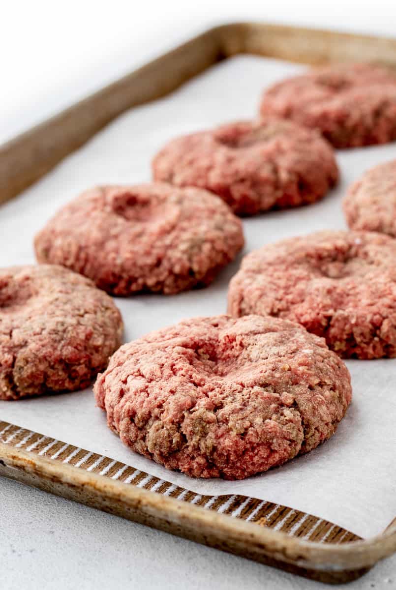 A baking sheet with parchment paper holding raw frozen burger patties with an indentation in the centre.