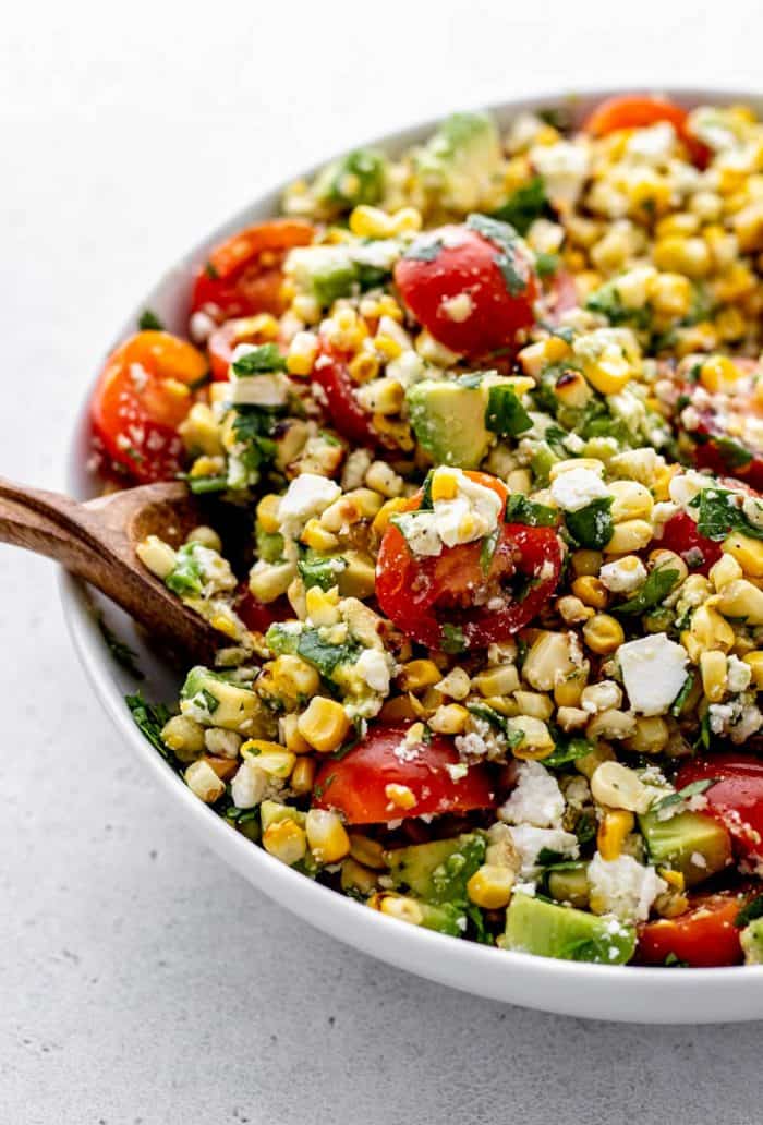 A wooden spoon digging into a bowl of grilled corn salad.
