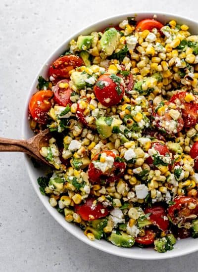A wooden spoon scooping up some corn avocado salad with feta, cilantro, and tomatoes.