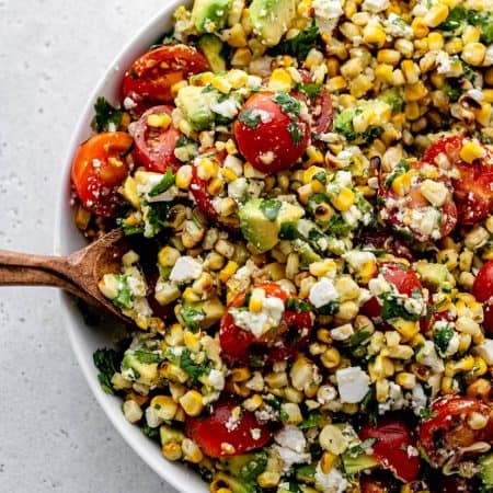 A wooden spoon scooping up some corn avocado salad with feta, cilantro, and tomatoes.