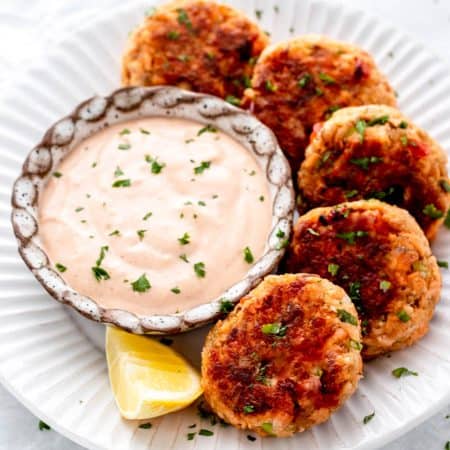 A white plate with salmon patties arranged around a bowl of dipping sauce with a lemon wedge on the side.
