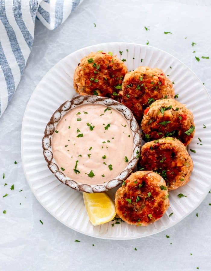 Overhead image of 5 healthy salmon patties topped with fresh herbs arranged around a bowl of creamy dipping sauce with a lemon wedge on the side.