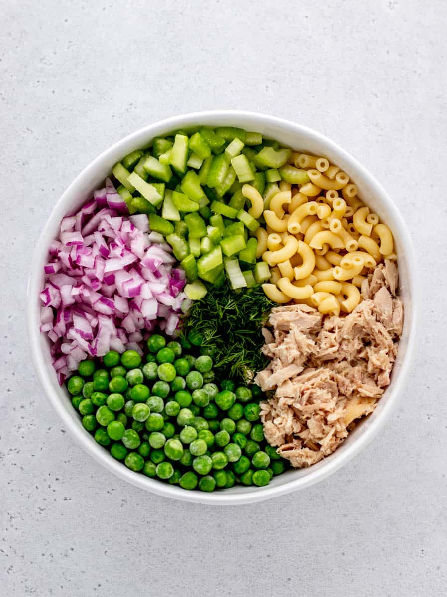 A white bowl full of chopped celery, red onions, frozen peas, flaked tuna, elbow macaroni, and fresh herbs.