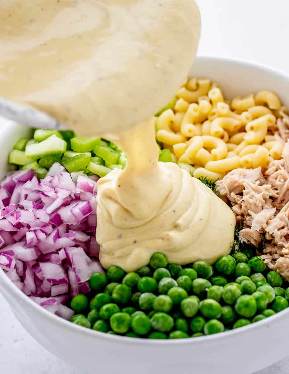 Greek yogurt dressing being poured on top of ingredients for tuna macaroni salad.