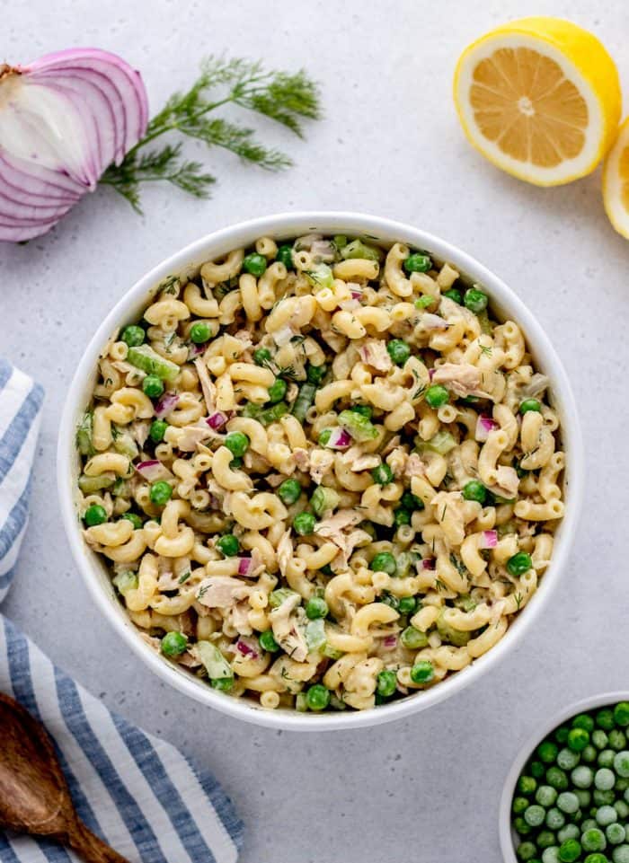 A white bowl full of tuna macaroni salad with half a red onion, fresh herbs, lemon halves, and frozen peas around the edges.