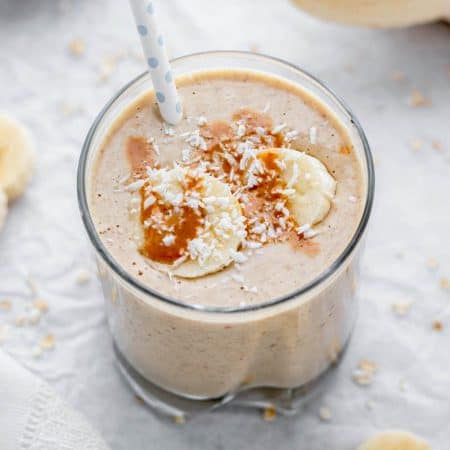 Creamy banana smoothie in a glass with a straw topped with peanut butter, coconut flakes and banana slices.