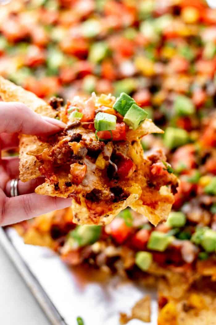 A hand holding up a loaded chip from a sheet pan of nachos supreme.