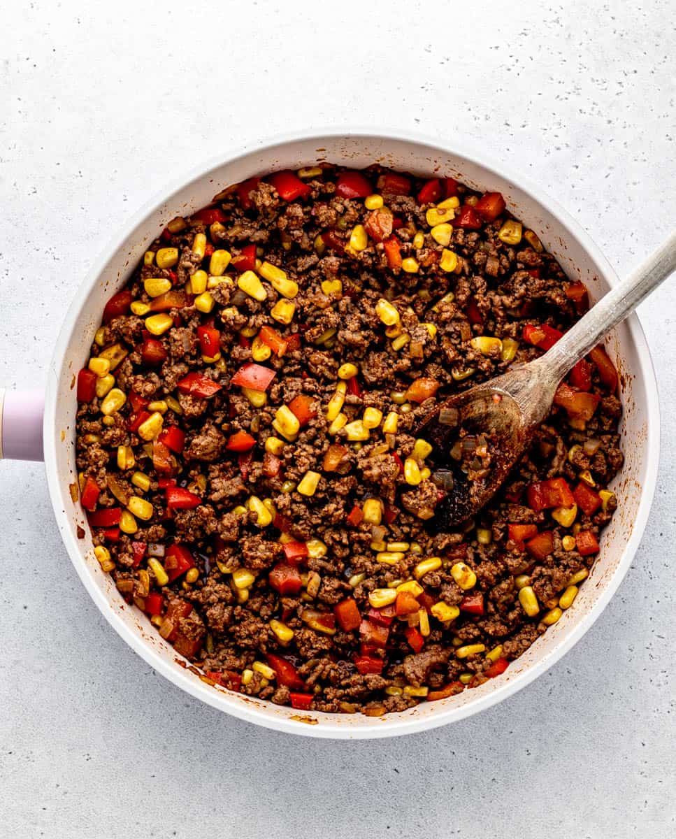 A skillet full of onions, peppers, lean ground beef, and corn being stirred with a wooden spoon.
