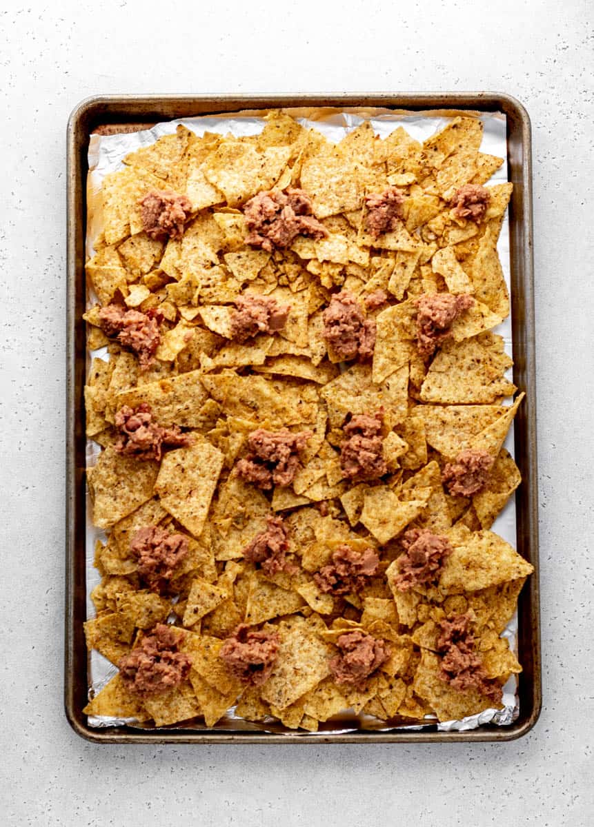 Nachos on a foil-lined baking sheet topped with refried beans.