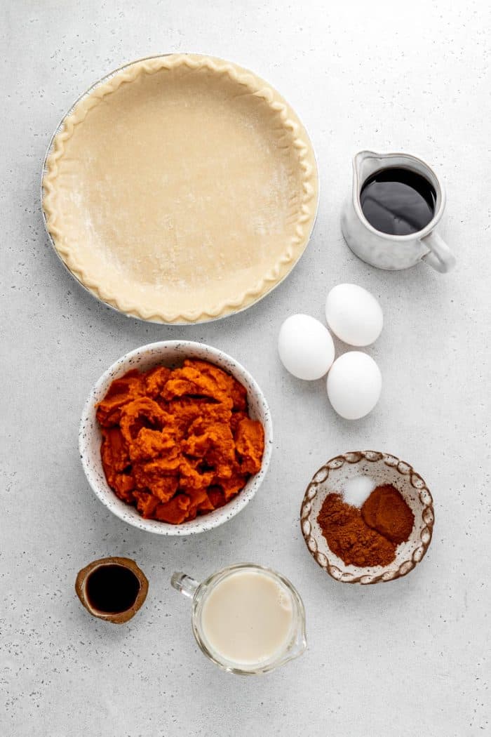 Overhead image of pie crust, three eggs, a bowl of pumpkin puree, spices, and maple syrup to make an easy pumpkin pie without evaporated milk.