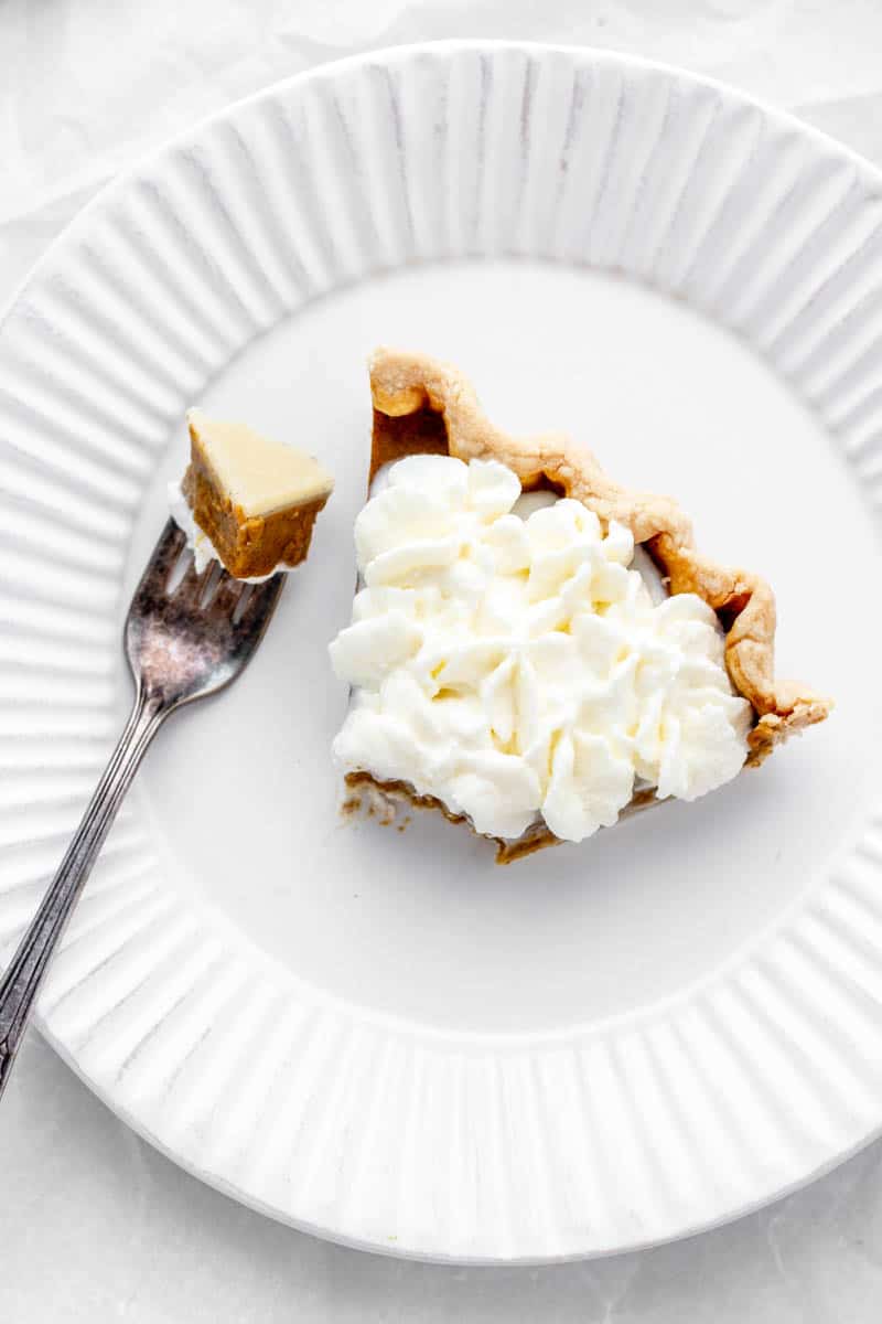 Overhead shot of a slice of pumpkin pie on a white plate topped with whipped cream.
