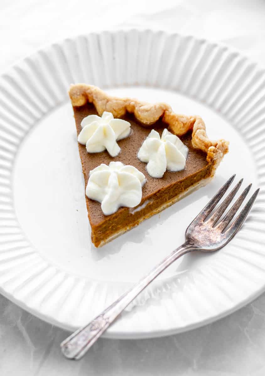 A slice of healthy pumpkin pie topped with three dollops of whipped cream on a white plate with a fork on the side.