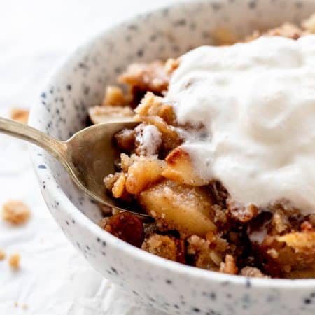 A spoon lifting a bite of healthy apple crisp topped with melting whipped cream.