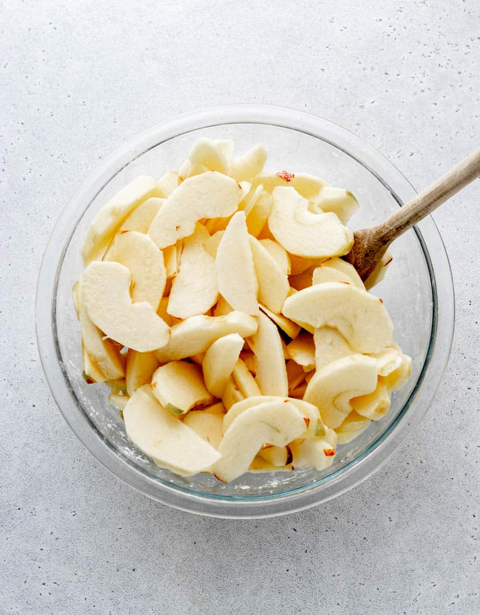 Overhead image of sliced apples in a glass bowl being stirred with a wooden spoon.