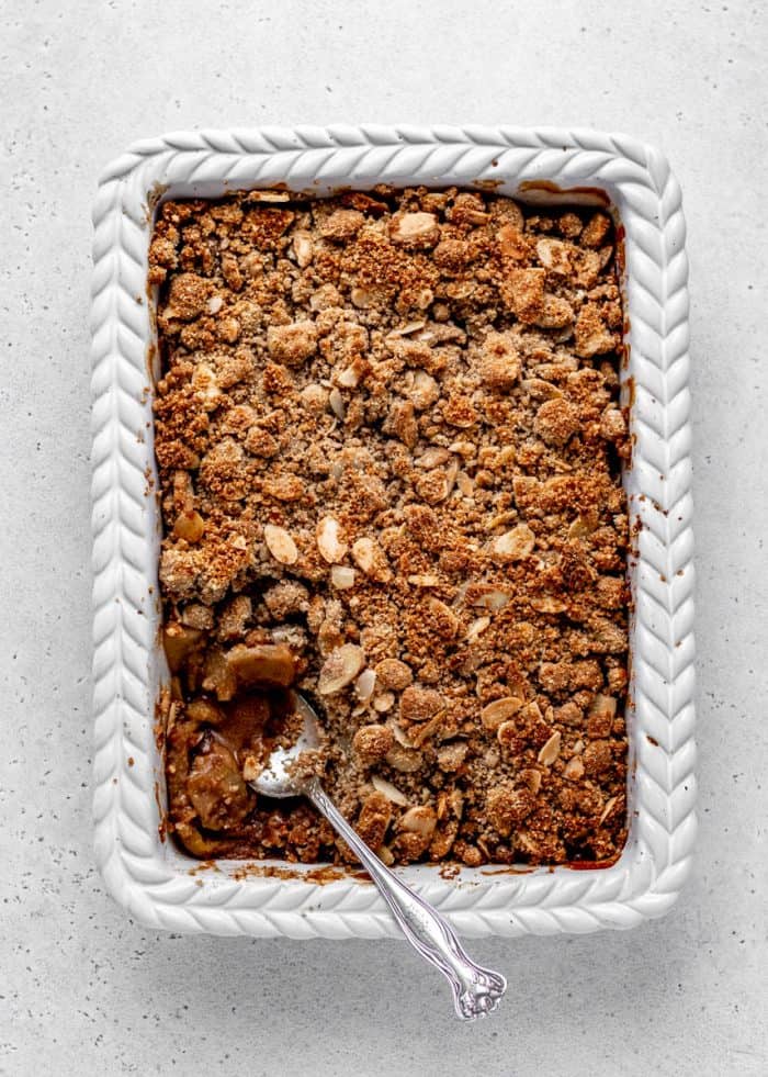 The baked apple crisp in a baking dish with a spoon digging into it.