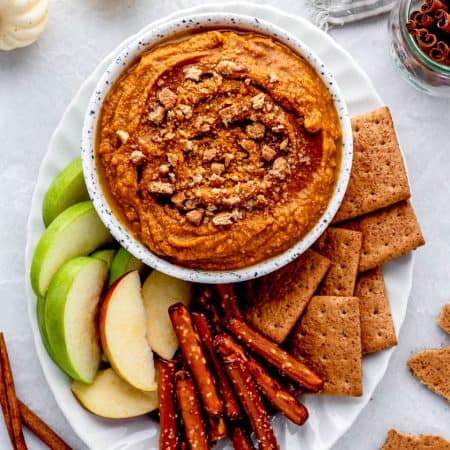 A white bowl full of pumpkin pie hummus topped with graham cracker crumbs on a white platter with graham crackers, pretzels, and green apples.