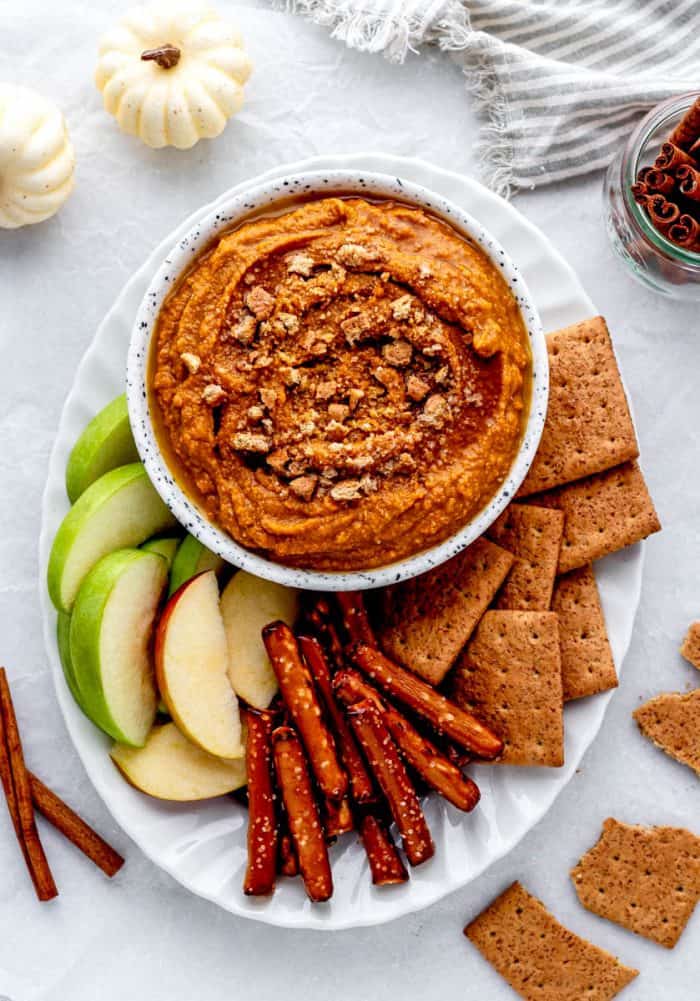 A white bowl full of pumpkin pie hummus topped with graham cracker crumbs on a white platter with graham crackers, pretzels, and green apples.